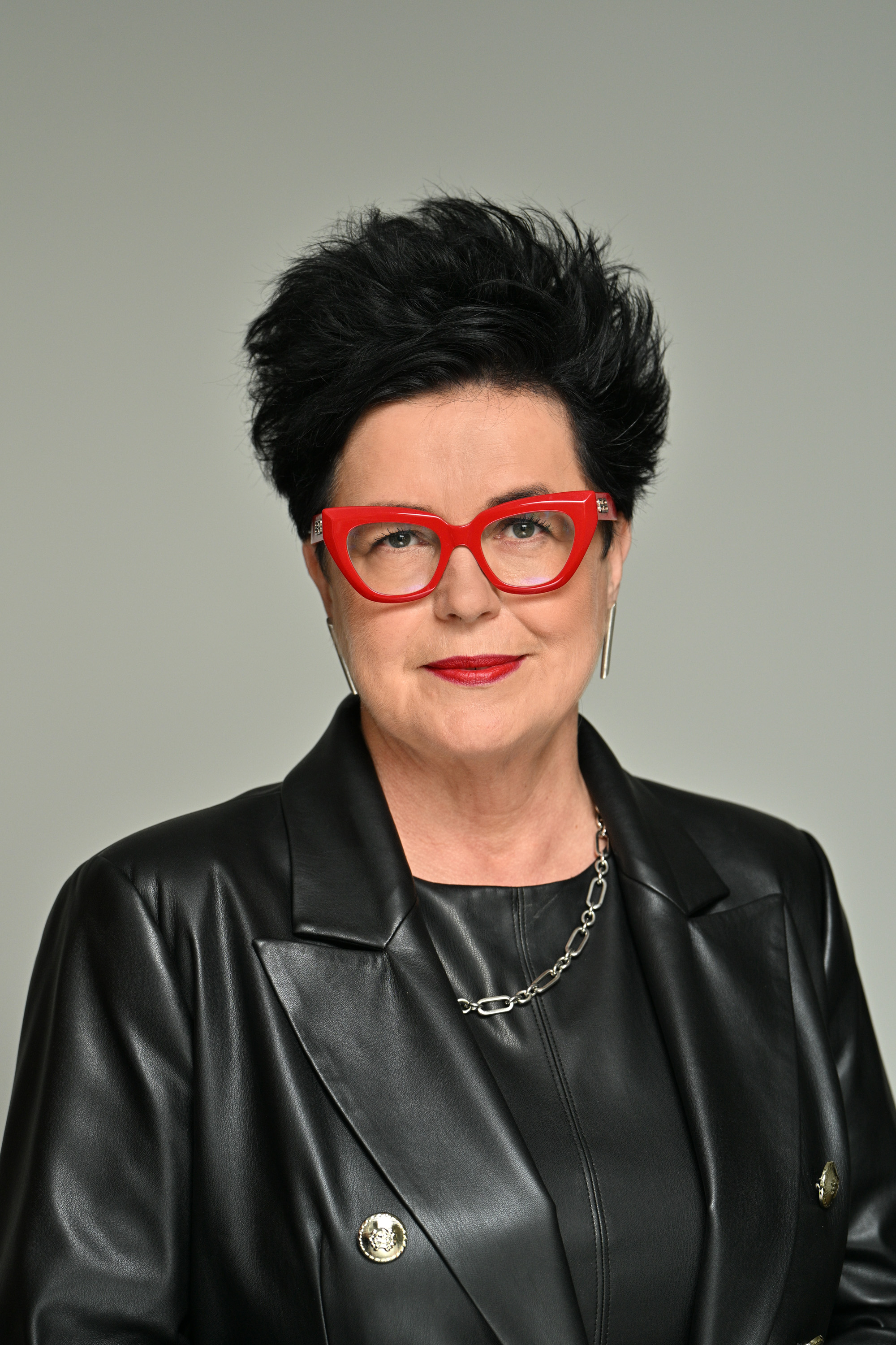 Fotografia prezentująca popiersie lekko uśmiechniętej, dojrzałej kobiety o ciemnych krótkich włosach. Ubrana jest w skórzaną marynarkę i top. Nosi okulary w grubych, czerwonych oprawkach, a na jej szyi widoczny jest srebrny łańcuszek o dużych ogniwach. Pozuje na jasno grafitowym tle.